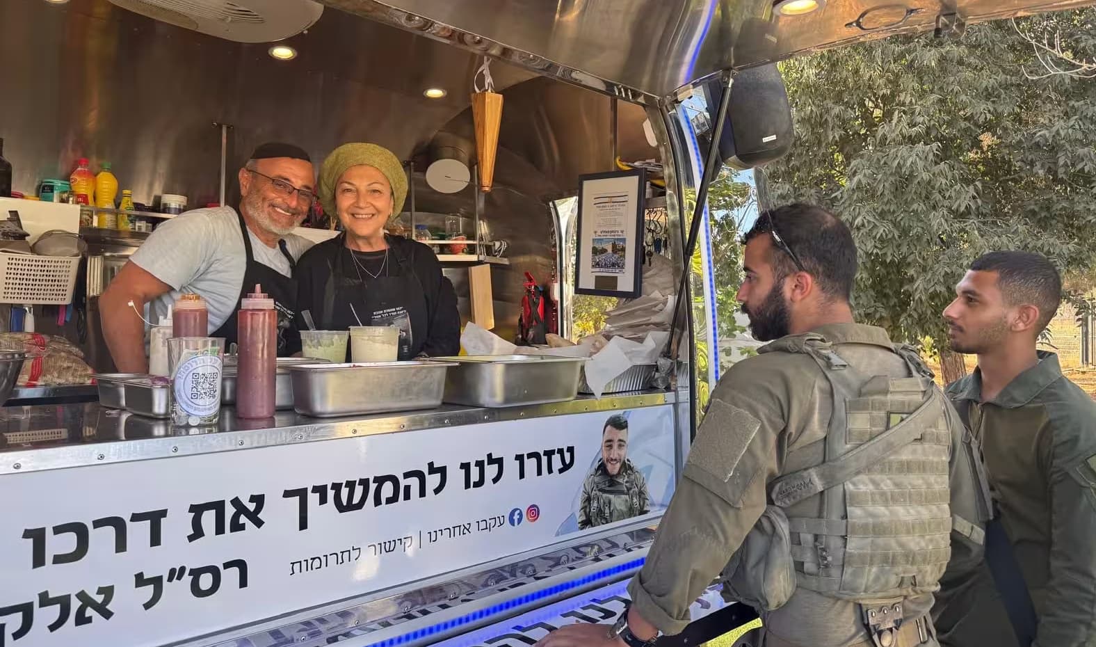 הפודטראק של יד אלקנה: גבר ואישה מחייכים מאחורי הדלפק, ושני חיילים עומדים מולם. על הדלפק שלט לזכר רס״ל אלקנה