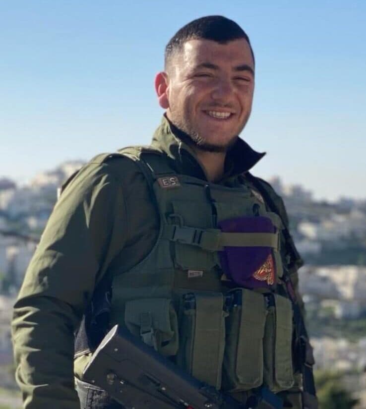 Elkana souriant, en uniforme et gilet de combat, un béret violet à l’épaule ; en arrière-plan, une ville sur une colline
