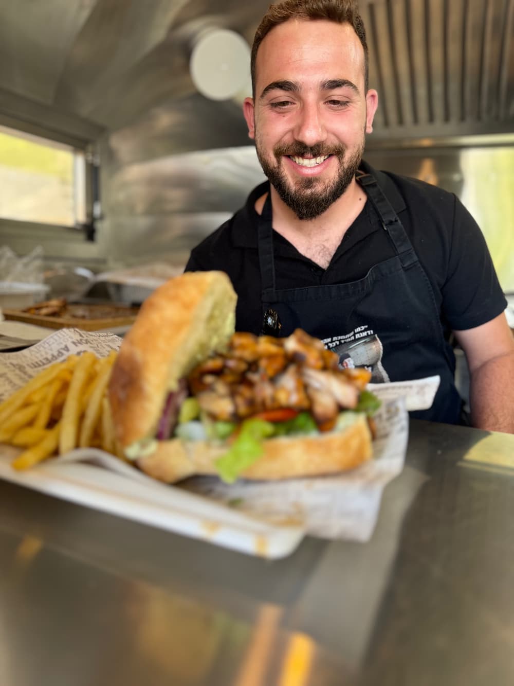 מתנדב מחייך בתוך הפודטראק מחזיק כריך גדול עם צ׳יפס