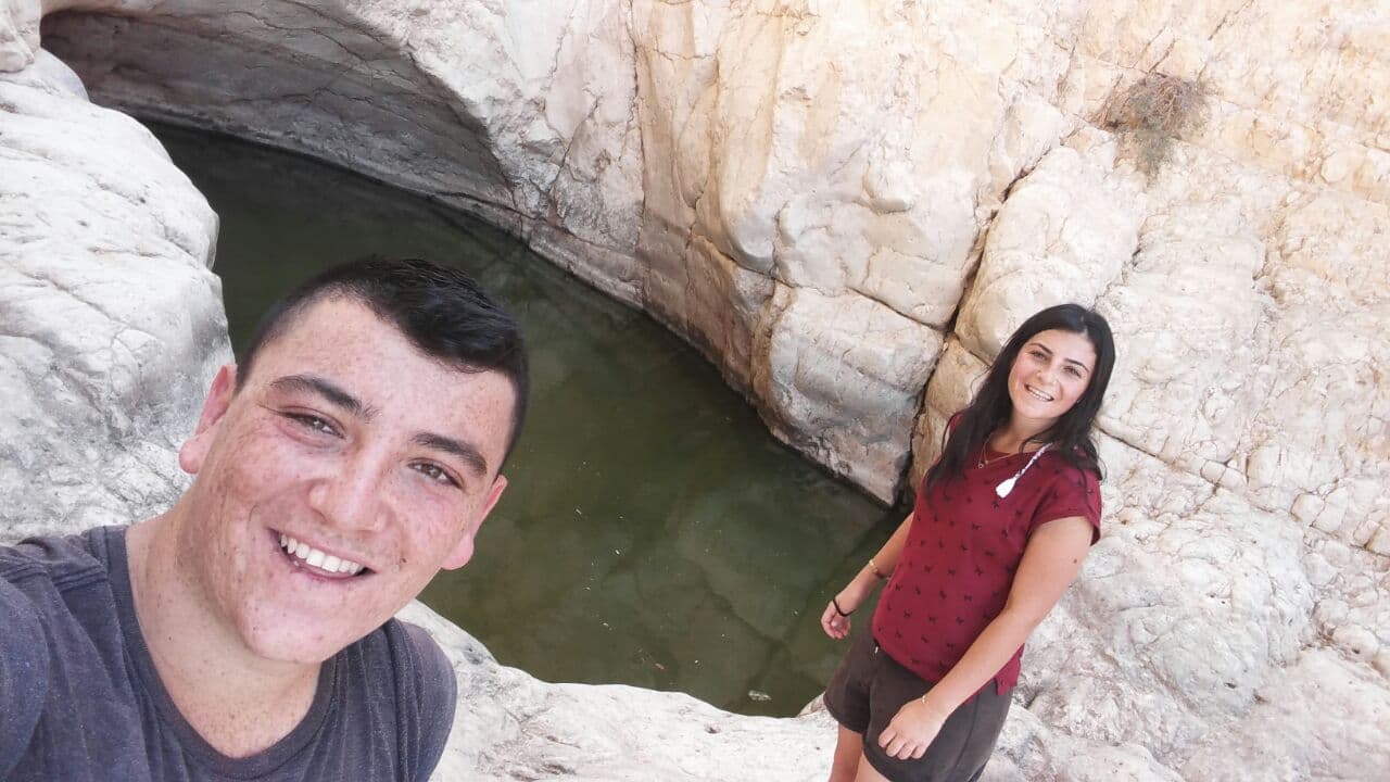 Elkana se prend en selfie, souriant, au bord d’un bassin entre les rochers, une jeune femme à ses côtés