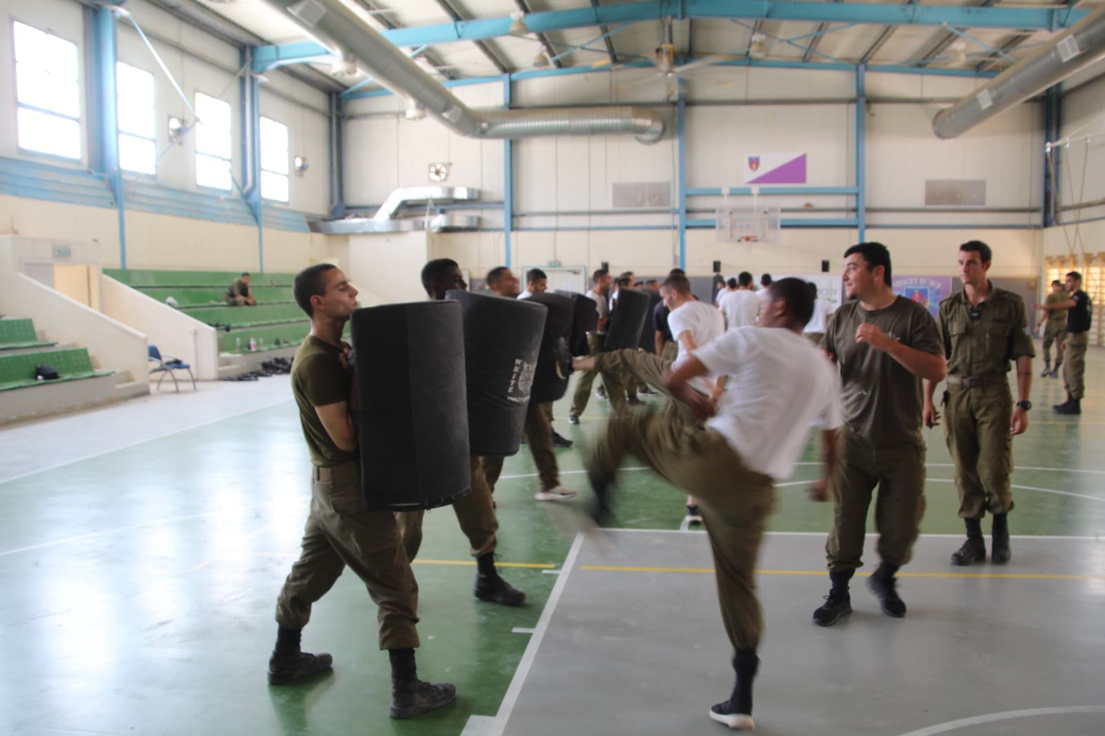 Des soldats à l’entraînement au combat rapproché dans un gymnase : l’un frappe du pied un bouclier tenu par un camarade