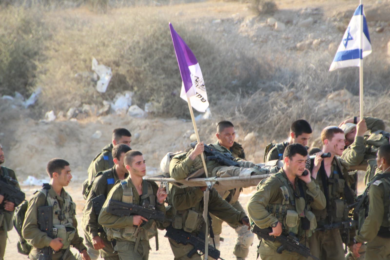 Des soldats portent un brancard lors d’une marche, sous un drapeau violet de Guivati et un drapeau israélien