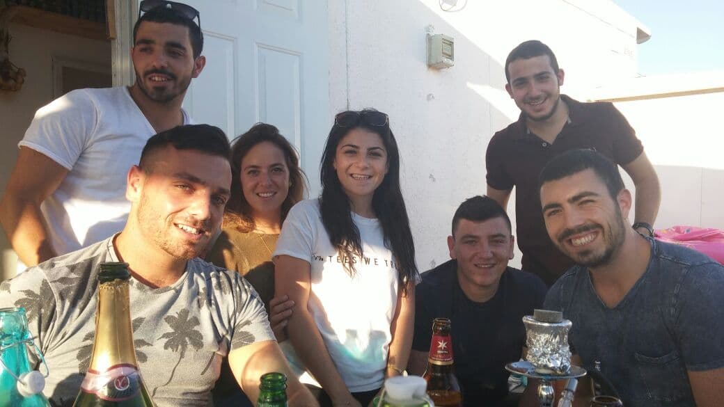 Sept jeunes gens, hommes et femmes, sourient autour d’une table en plein air