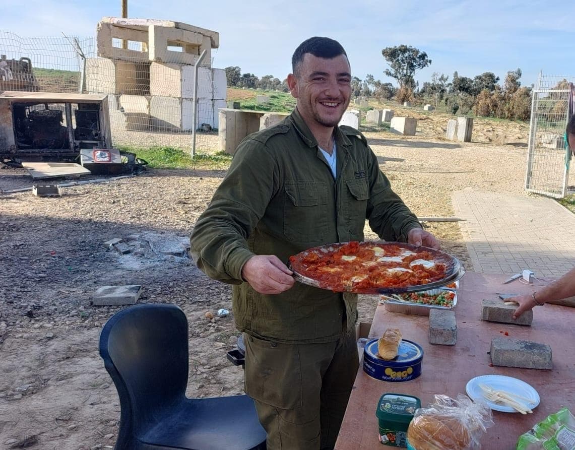 Elkana en uniforme, souriant, sert une grande poêle de chakchouka près d’une table, sur le terrain