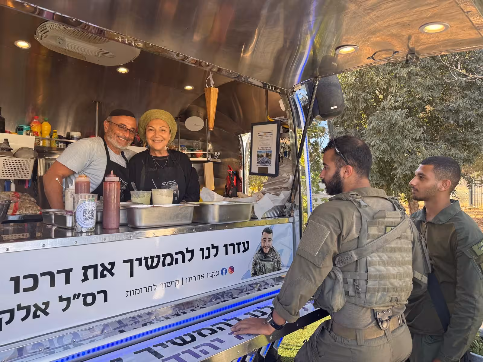 Le food-truck de Yad Elkana : un homme et une femme sourient derrière le comptoir, deux soldats se tiennent devant. Une banderole sur le comptoir rend hommage à Elkana